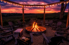 A circular fire pit surrounded by wooden chairs and tables, with string lights overhead.