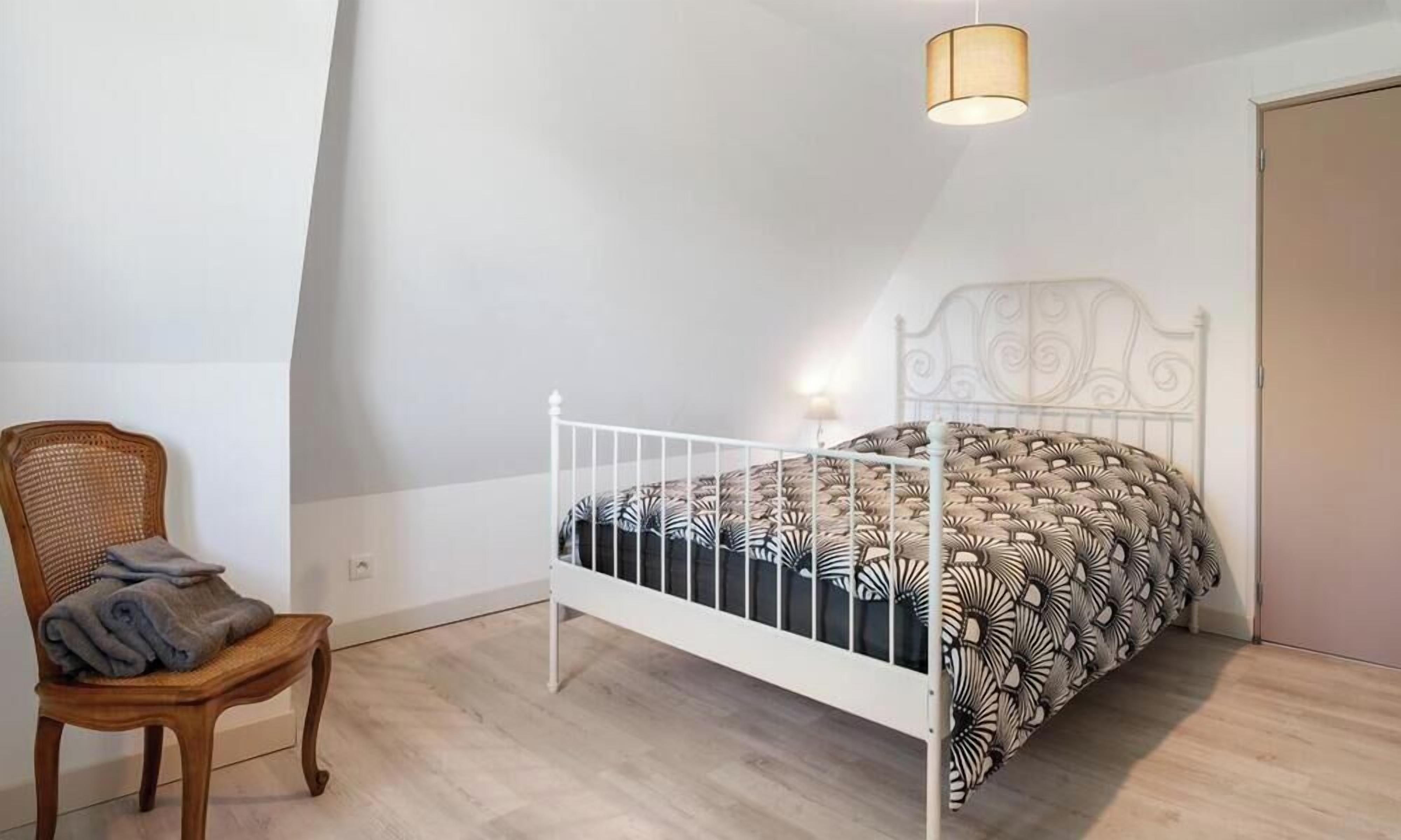 Een slaapkamer met een bed van wit metaal, een houten stoel met kussen, en een wandlamp.