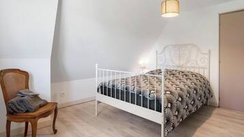 Een slaapkamer met een bed van wit metaal, een houten stoel met kussen, en een wandlamp.