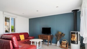 A modern living room with a red leather sofa, a white table, a television on a wooden stand, and a wood-burning stove. - Gîtes de France® - Le Noyer Gourmand (ILLOIS)
