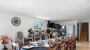 A modern kitchen with a dining area, wooden chairs, a clock, and a staircase. - Gîtes de France® - Le Noyer Gourmand (ILLOIS)