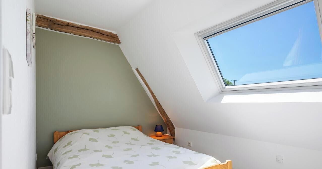 Una habitación pequeña con cama individual, techo inclinado, ventana y mesita de noche con lámpara.