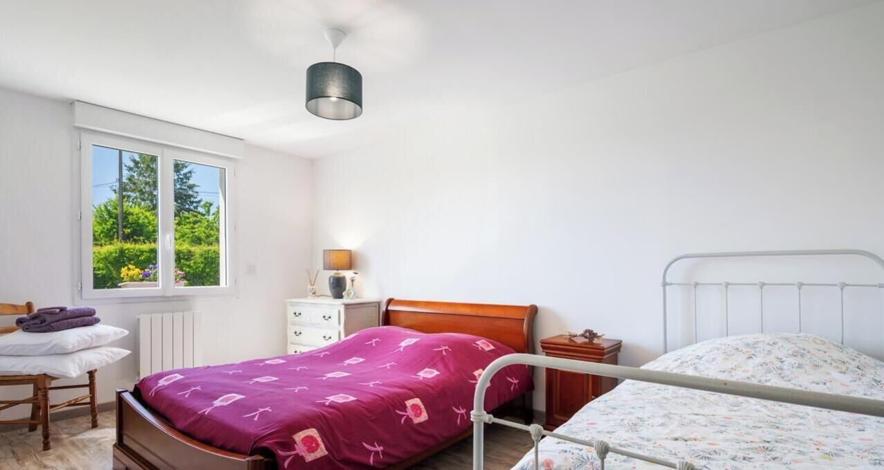A bedroom with two beds, a window with a view of greenery, a bedside table with a lamp, and a radiator.
