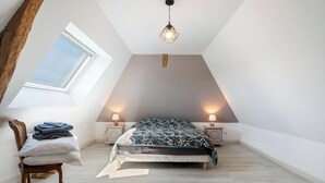 A bedroom with a sloped ceiling, a skylight, a bed with a patterned bedspread, two bedside lamps, and a chair with folded clothes. - Gîtes de France® - Le Noyer Gourmand (ILLOIS)