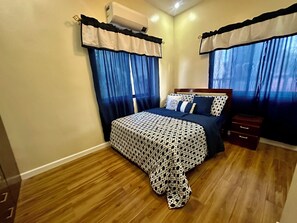 A bedroom with a bed, curtains, a nightstand, and an air conditioning unit. - BethShalom Guest House by Jeff and Malou, 4 Bedrooms and 2 Bathrooms (Dasmarinas)