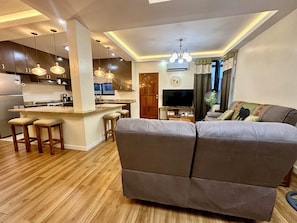 A modern living room with a grey sofa, wooden flooring, and a kitchen area with dark cabinets and pendant lighting. - BethShalom Guest House by Jeff and Malou, 4 Bedrooms and 2 Bathrooms (Dasmarinas)