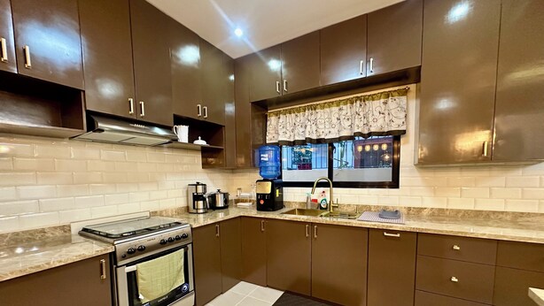 A modern kitchen with dark brown cabinets, a stainless steel stove, and a window with a decorative curtain. - BethShalom Guest House by Jeff and Malou, 4 Bedrooms and 2 Bathrooms (Dasmarinas)