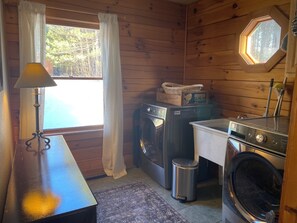 A compact kitchen with wooden walls, a washing machine, a dryer, a trash bin, and a window with curtains.