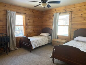 A room with wooden walls, two single beds, a ceiling fan, and a window with curtains.