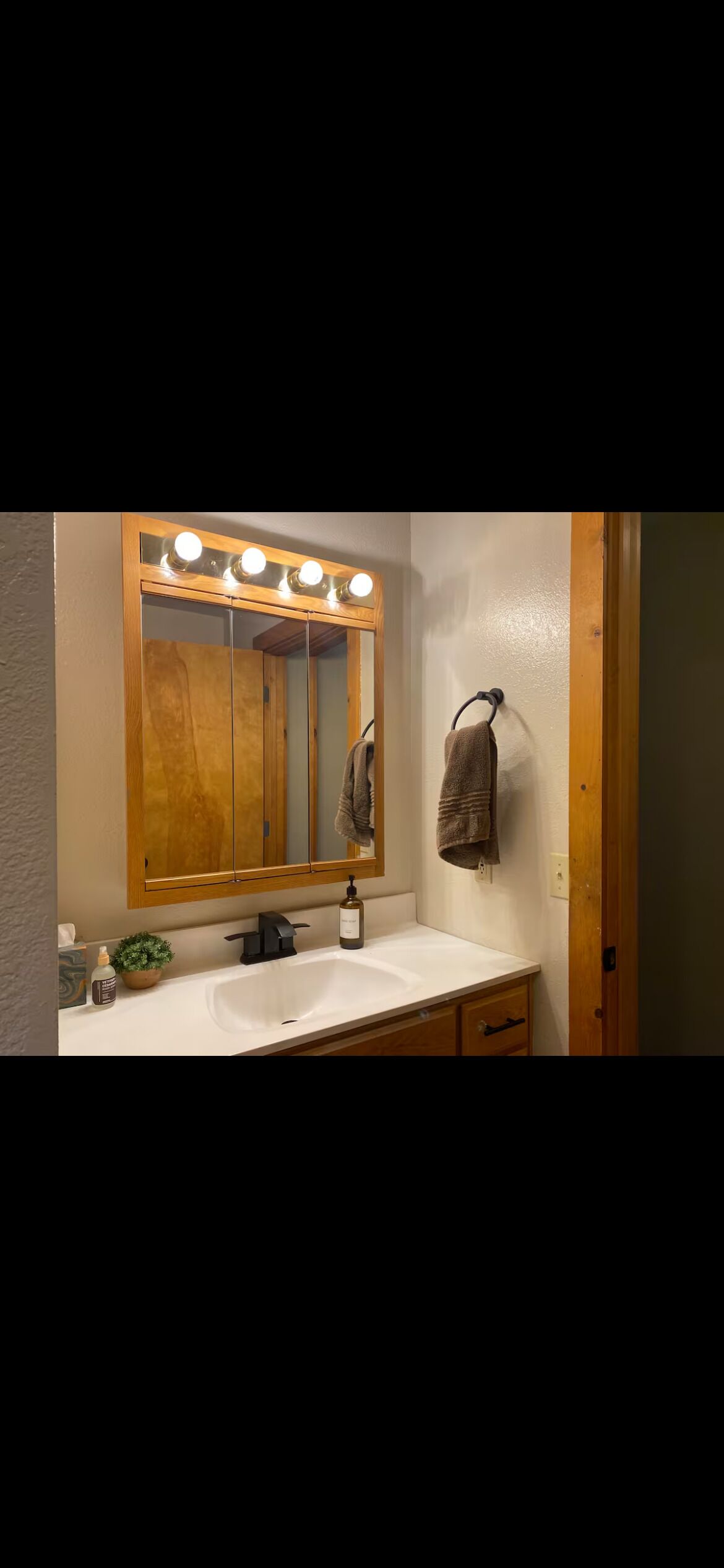 A bathroom with a wooden vanity, a mirror with lights, a towel rack, and a small plant.
