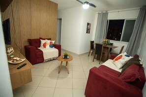 Ein Wohnzimmer mit einem roten Sofa, einem kleinen runden Tisch mit einer Pflanze, einem Esstisch mit Stühlen und einem Fenster mit Vorhängen.