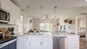 A modern kitchen with white cabinets, a dishwasher, a microwave, and a coffee maker. There is a dining area with a table and chairs, and a bedroom with a bed and a nightstand. - Palm Springs Oasis w/Pool & Golf Access (Cathedral City)