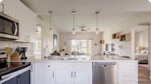 Eine moderne Küche mit weißen Schränken, einem Geschirrspüler, einer Mikrowelle und einer Kaffeemaschine. Es gibt einen Essbereich mit Tisch und Stühlen sowie ein Schlafzimmer mit Bett und Nachttisch.