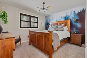 A bedroom with a wooden bed, a desk, a chair, a window, and a wall mural.