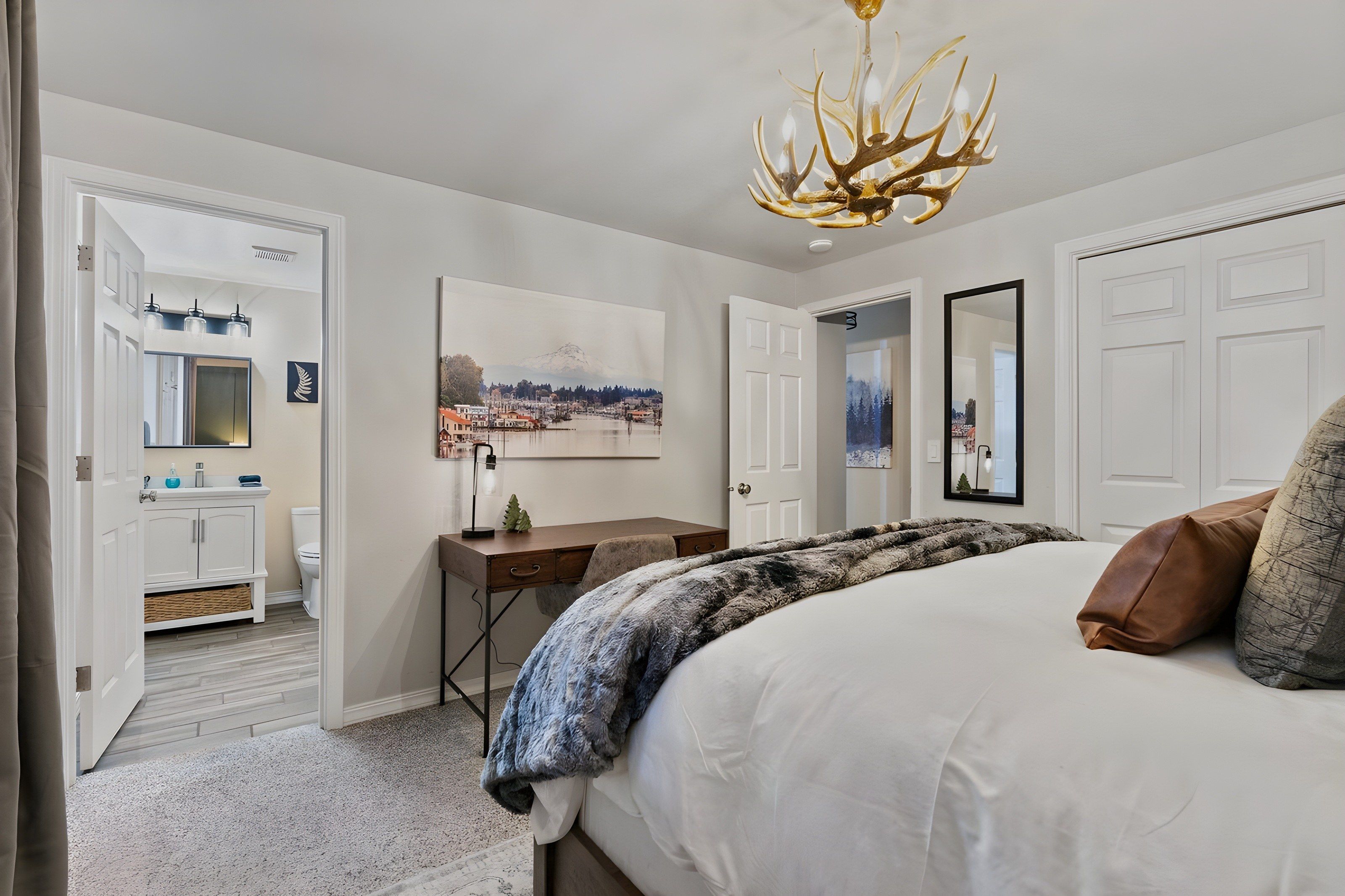 A bedroom with a large bed, a desk, a chair, a mirror, and a painting of a mountain landscape.