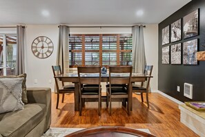 Un coin repas avec une table et des chaises en bois, une grande horloge murale et une fenêtre avec des stores.
