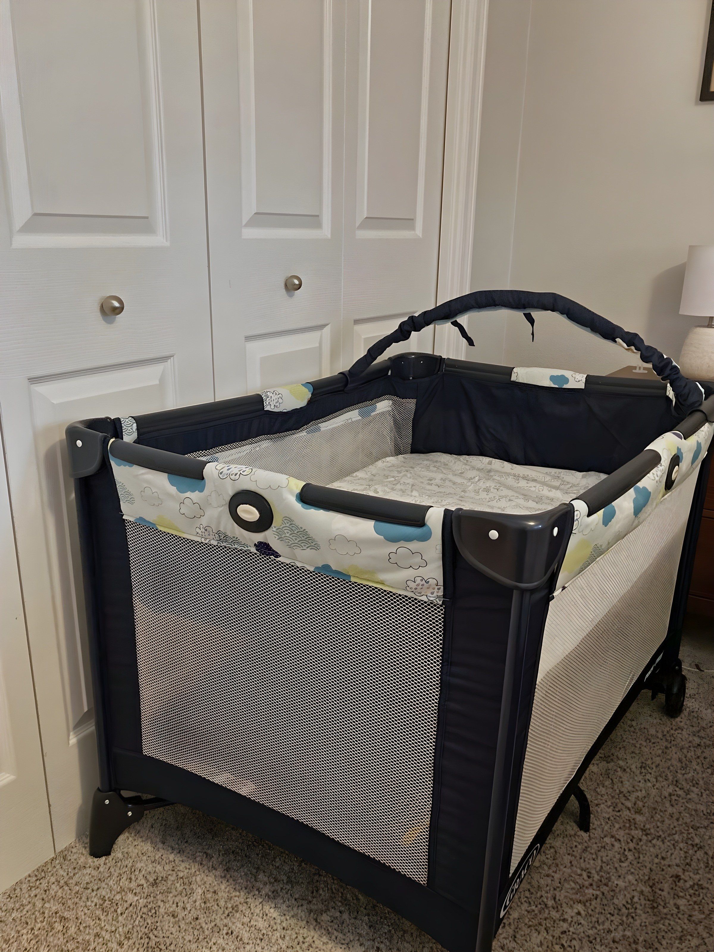 A portable crib with a mesh front and a handle, placed against a wall with a door and a lamp.
