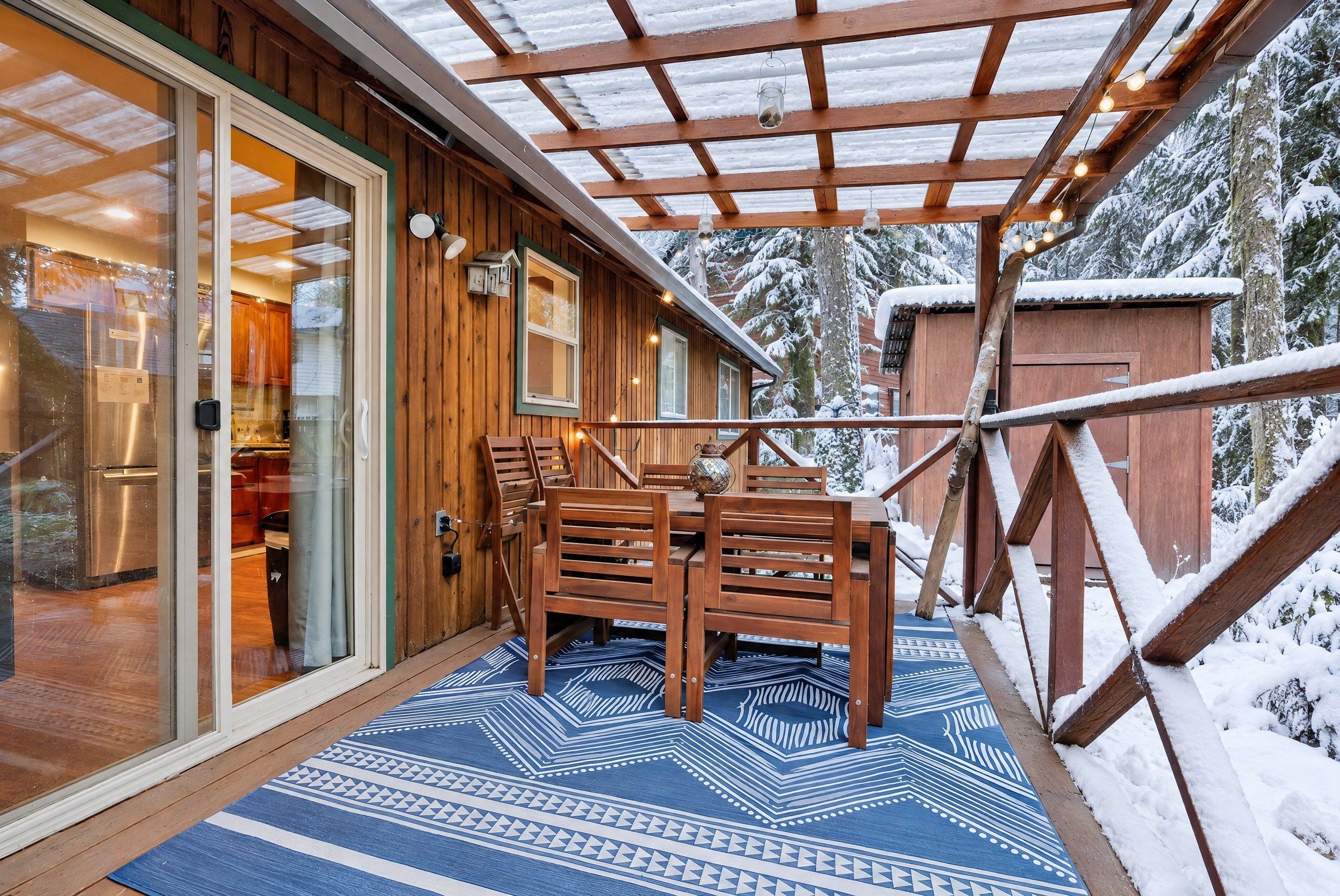 A wooden exterior with a covered patio area, a dining table with chairs, and a snow-covered landscape.