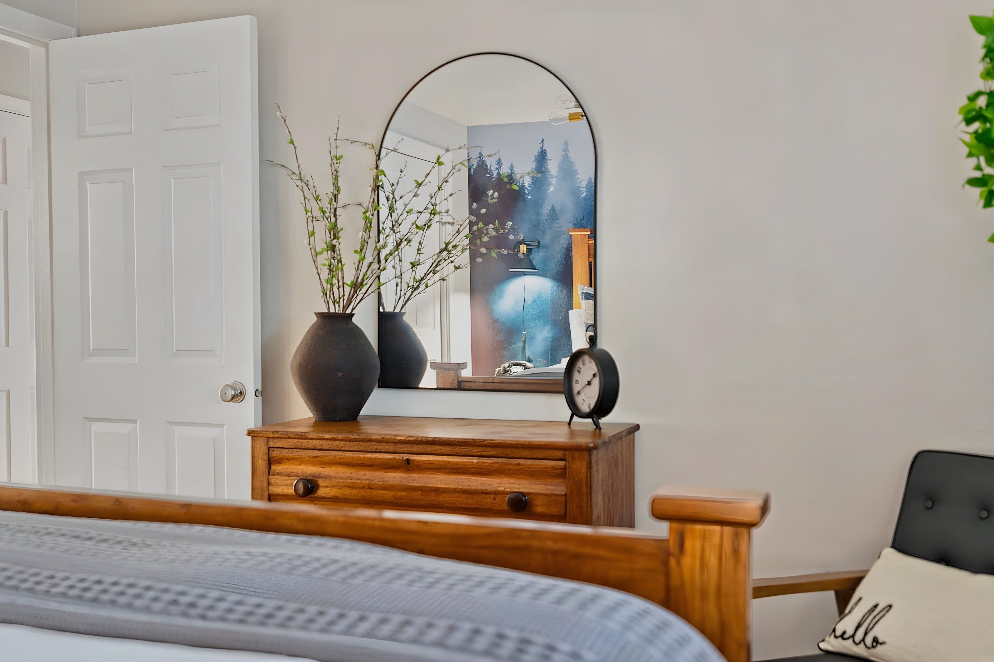 Ein Schlafzimmer mit einem Holzkoffer, einem Spiegel, einer Uhr und einem Bett mit grauem Kopfteil.
