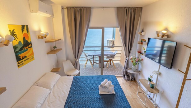 Ett hotellrum med en säng, en TV, en balkong med ett bord och stolar samt utsikt över havet.