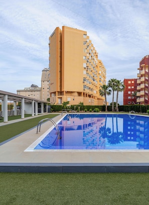 Una piscina amb una superfície d'aigua blava i cristal·lina, envoltada d'un jardí ben cuidat i un edifici alt amb múltiples balcons al fons.