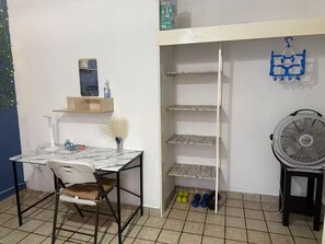 Une pièce comprenant un bureau, une chaise, des étagères, un ventilateur et du matériel d’exercice.