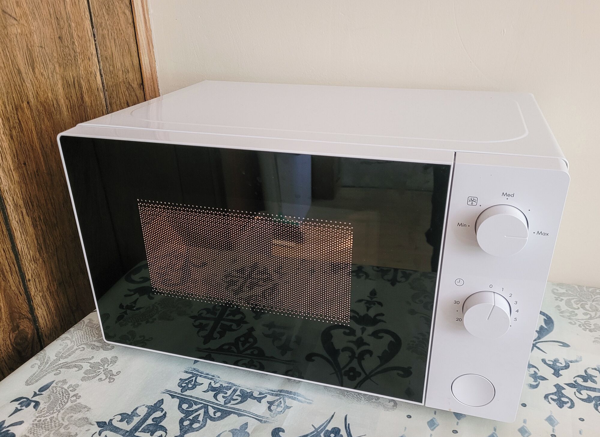 Un microondas blanco con interior negro, un panel de control con temporizador y ajustes de temperatura, y un mantel estampado debajo.