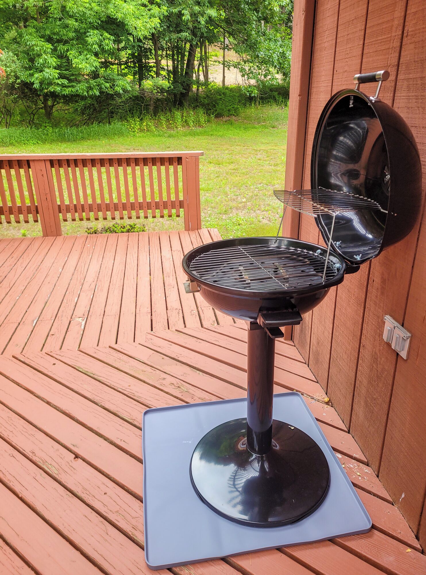 A portable grill on a wooden deck with a view of a grassy area and trees.