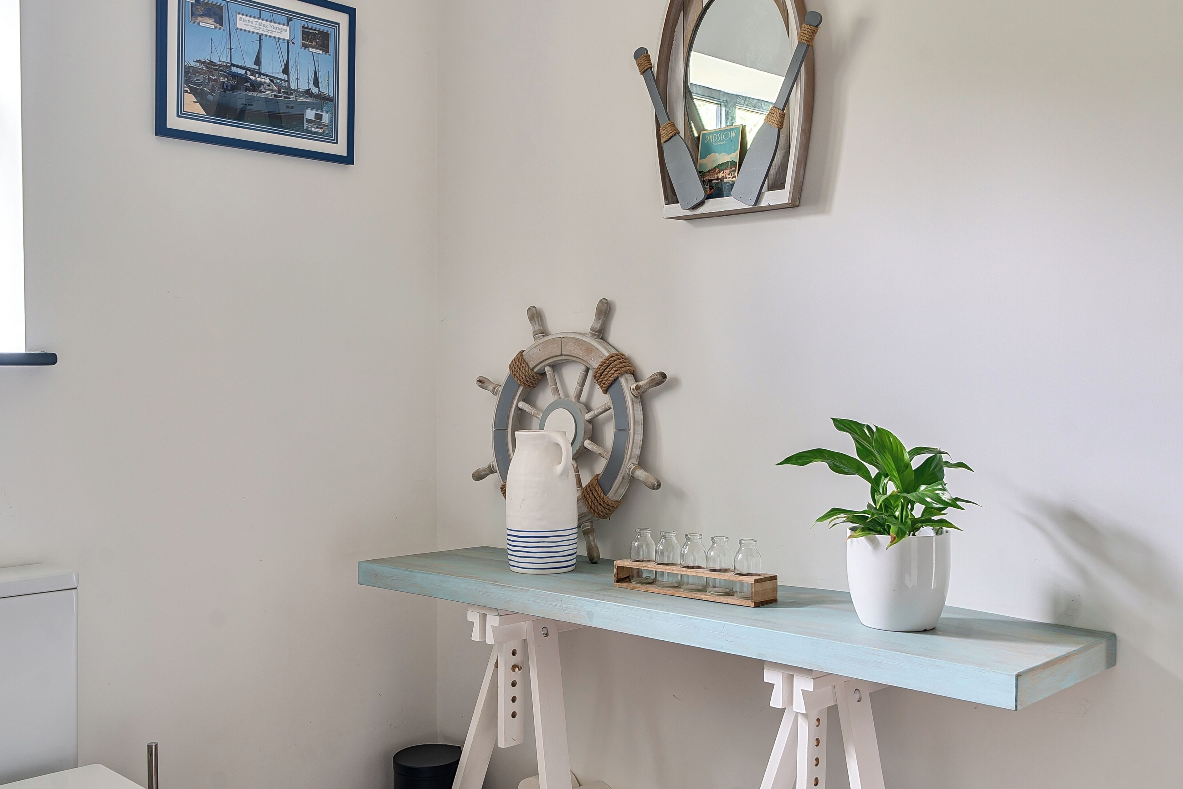 Ein Zimmer mit hellblauer Wand, einer hölzernen Schiffslenkerrade als Dekoration, einem gerahmten Bild, einer Topfpflanze und einem Regal mit Gläsern.