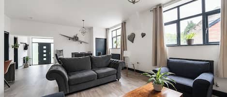 Een ruime woonkamer met een grijze bank, een houten salontafel en een groot raam met gordijnen.