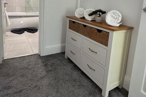 Et badeværelse med et hvidt badekar, et hvidt toiletbord med fletkurve og et gråt tæppedækket gulv.