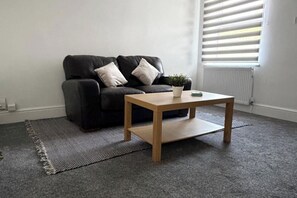 En stue med en sort sofa, et træbord, en lille potteplante og en radiator.