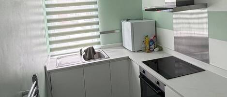 A compact kitchen with a built-in oven, a small refrigerator, and a window with blinds.