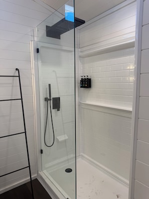 A glass-enclosed shower with a black towel rack, a glass shelf with toiletries, and a white tiled wall.