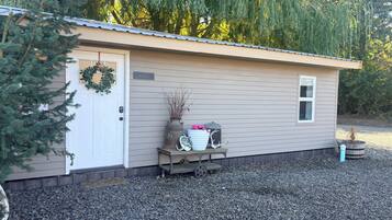 A small, single-story building with a door and a window, surrounded by gravel and a weeping willow tree.