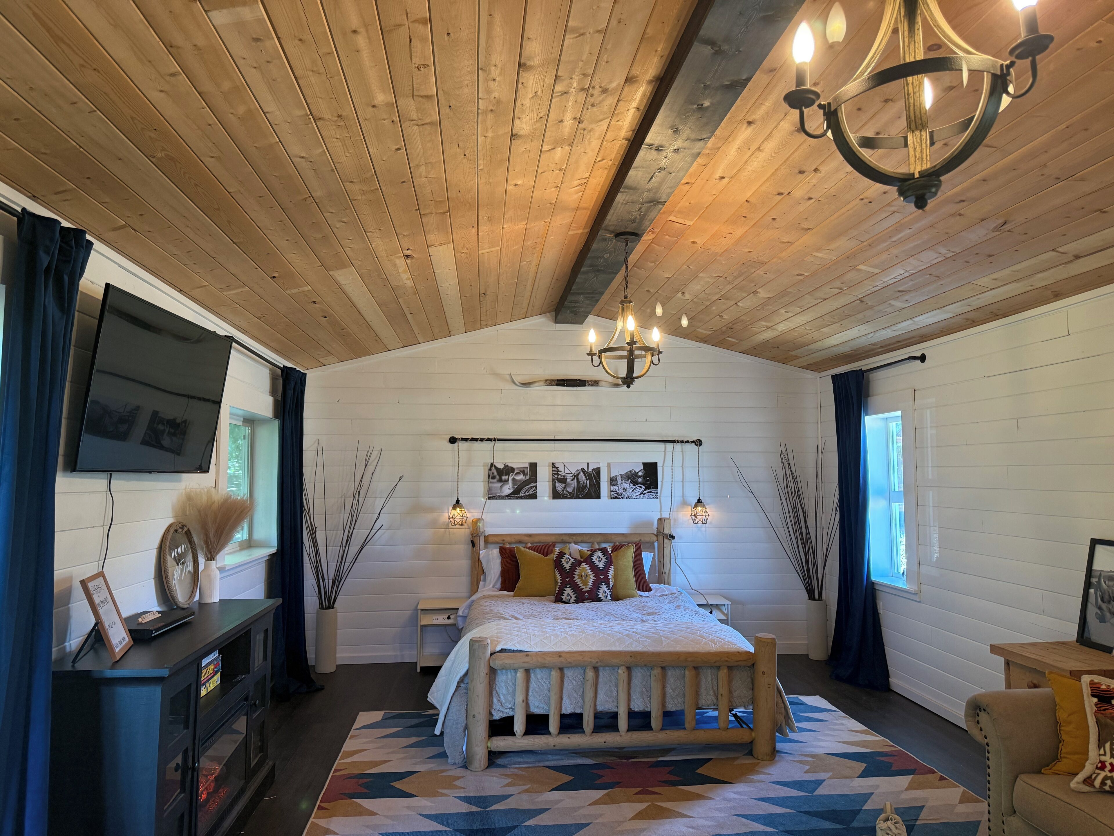 A bedroom with a wooden ceiling, a bed with a patterned bedspread, a chandelier, and a television on the wall.. Willow Pond Cabin