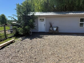 A gravel driveway leading to a white building with a metal roof, a wooden fence, and a large tree. - Willow Pond Cabin (Ellensburg)
