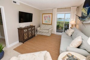 Uma sala de estar com sofá, uma poltrona estampada, um armário de madeira, uma televisão e vista para uma varanda com corrimão.