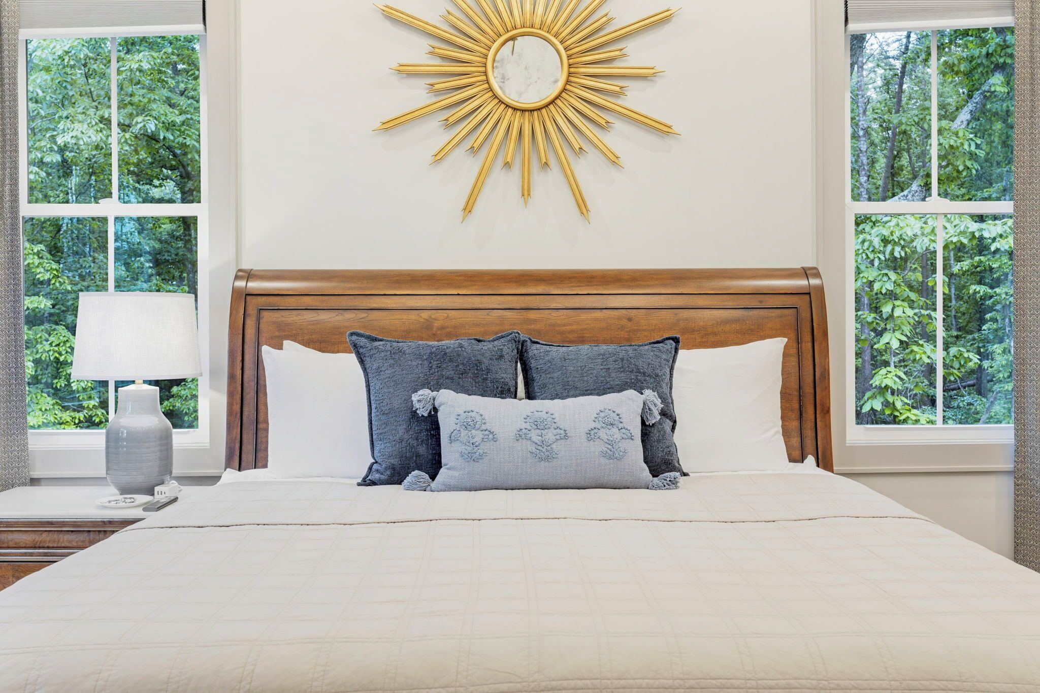 Une chambre à coucher avec une tête de lit en bois, un lit recouvert d’une couette blanche et grise, et un miroir en forme de soleil accroché au mur.