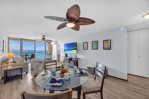 A modern living room with a glass dining table, a ceiling fan, and a large TV. - Oceanfront 2 Bedroom Oasis on Sugar Beach (Wailuku)