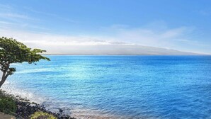 Vista costeira com céu claro, mar calmo e cadeia montanhosa ao longe.