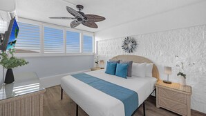 Um quarto com cama, ventoinha no teto, janela com persianas, objeto decorativo na parede e mesa de cabeceira com abajur.