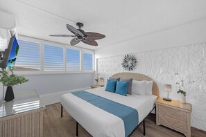 Ein Schlafzimmer mit einem Bett, einem Deckenventilator, einem Fenster mit Jalousien, einem Wanddekor und einem Nachttisch mit Lampe.