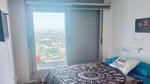 A bedroom with a large window, a bed with a patterned bedspread, a wall-mounted TV, and a ceiling light. - Apartment on Shopping (Montevideo)