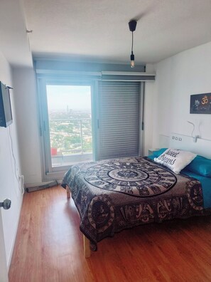 Un dormitorio con un ventanal grande, una cama con colcha estampada, un televisor montado en la pared y una lámpara del techo.