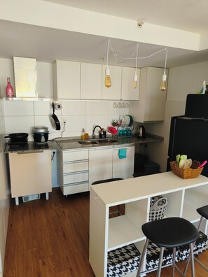 Una cocina con armarios blancos, un refrigerador negro, una encimera con un hervidor y una zona de comedor con taburetes.
