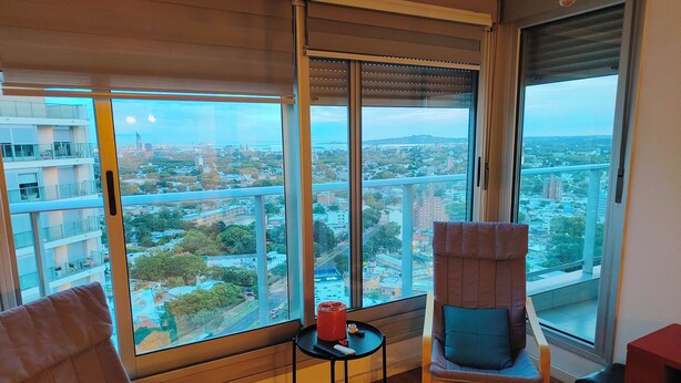 A room with a large window, two chairs, a small table, and a red object on the table. - Apartment on Shopping (Montevideo)