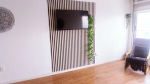A modern living room with a flat-screen TV mounted on a wall with vertical stripes, a wooden floor, a grey sofa, and a window with natural light. - Apartment on Shopping (Montevideo)
