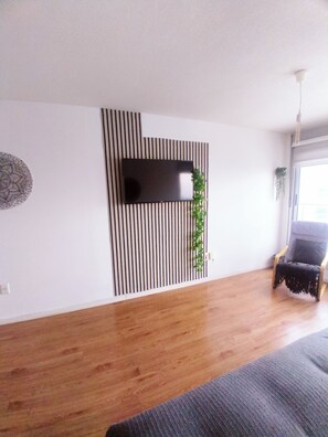 Un salón moderno con un televisor de pantalla plana montado en una pared de rayas verticales, suelo de madera, un sofá gris y una ventana con luz natural.