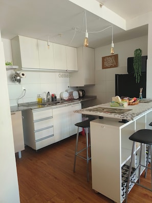 Una cocina moderna con armarios blancos, una isla central con taburetes, una lámpara colgante y un letrero en la pared.